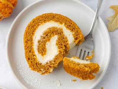 A slice of pumpkin roll on a white plate with a fork taking a bite.