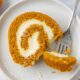 A slice of pumpkin roll on a white plate with a fork taking a bite.