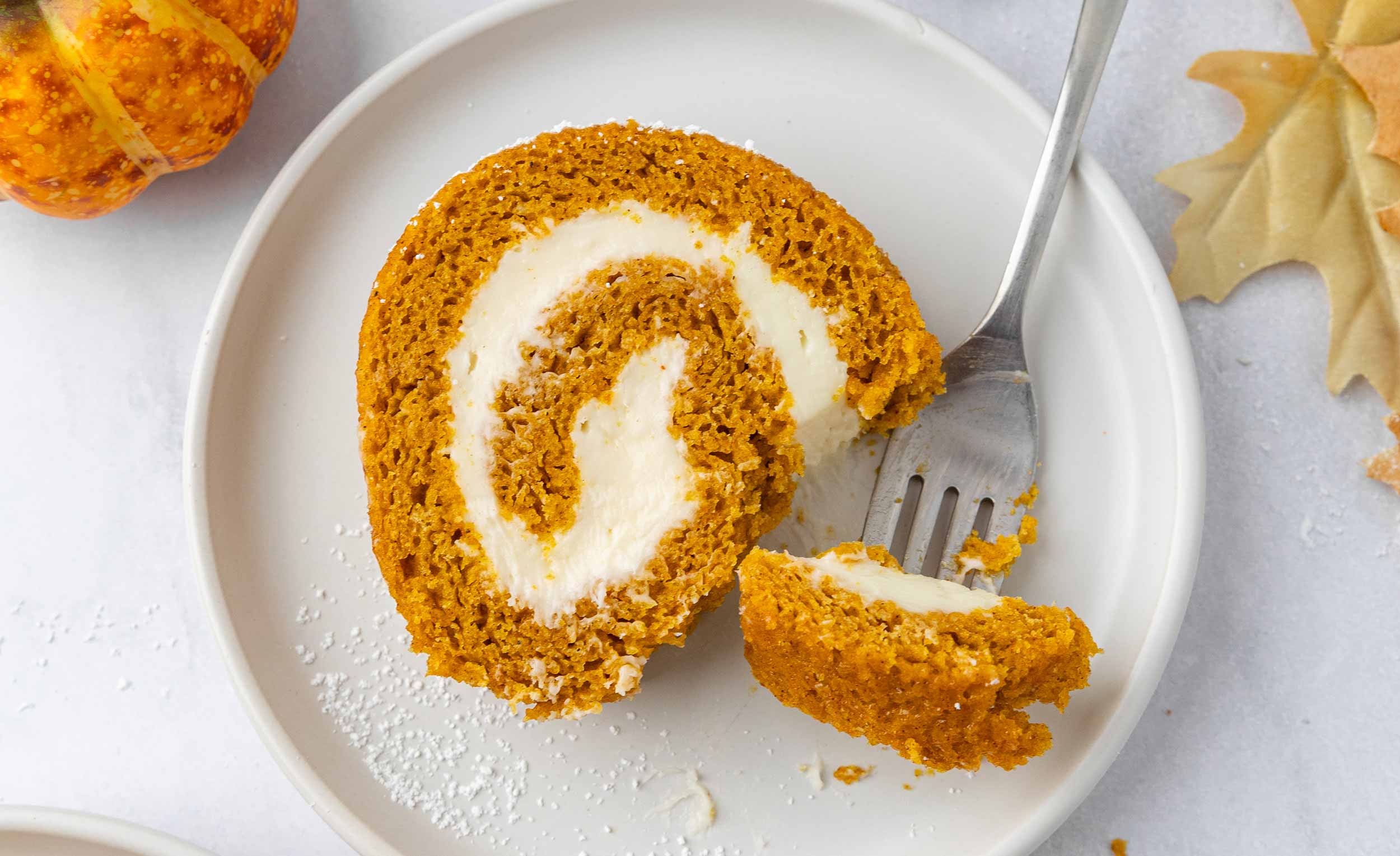 A slice of pumpkin roll on a white plate with a fork taking a bite.