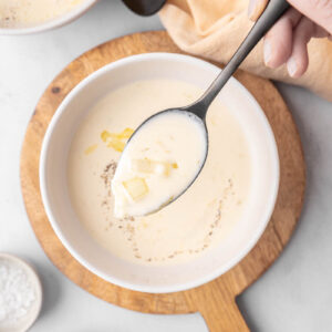 Cream of onion soup on a spoon with a white bowl of soup on a wooden cutting board.