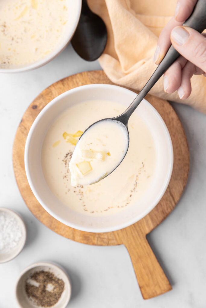 Cream of onion soup on a spoon with a white bowl of soup on a wooden cutting board.