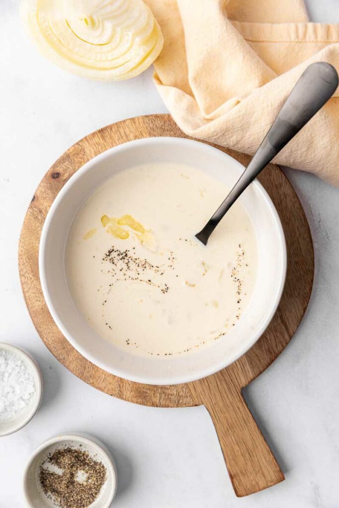 Cream of onion soup on a spoon with a white bowl of soup on a wooden cutting board.