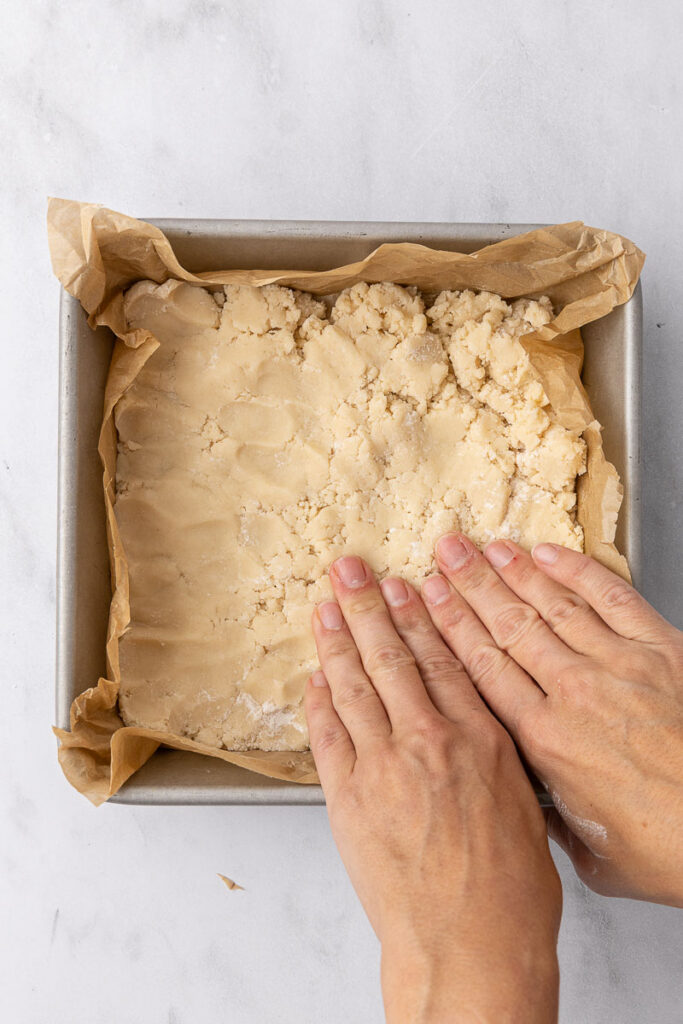 Hands pressing a cookie crust into a square parchment paper lined pan.