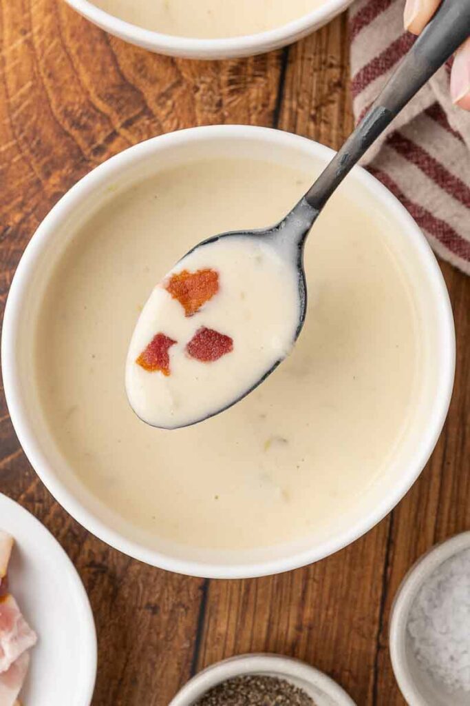 A spoonful of cream of bacon soup with soup in white bowls on a wooden counter.