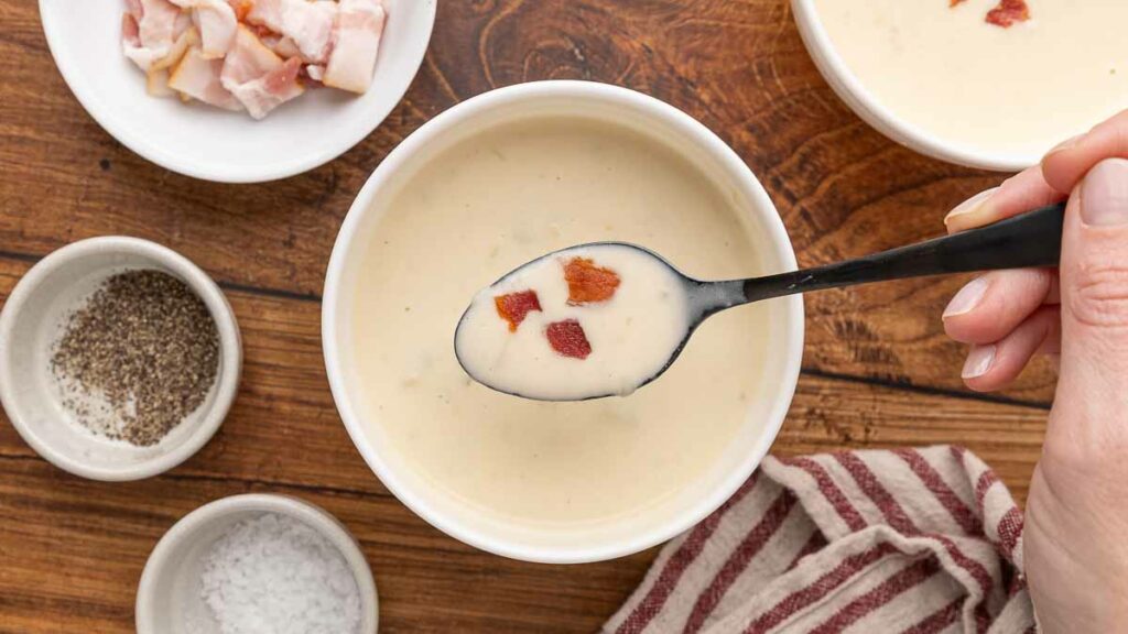 A spoonful of cream of bacon soup with soup in white bowls on a wooden counter.