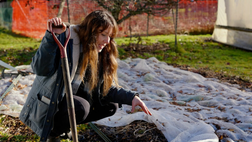 A woman holding a shovel crouched by a garden in the winter.