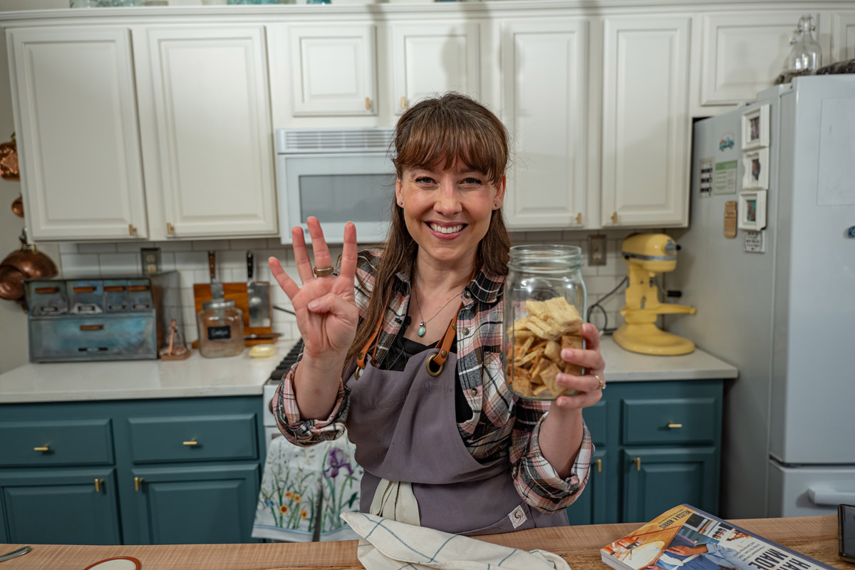 A woman holding a jar of homemade crackers and holding up four fingers to indicate this recipe only needs four ingredients.