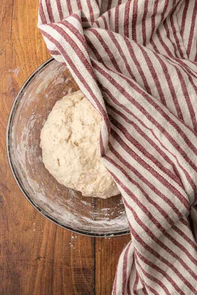 A ball of no-knead bread dough sitting in a glass bowl getting covered with a tea towel.