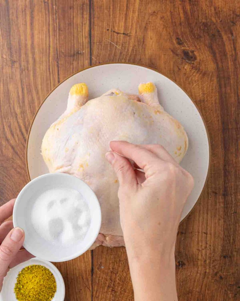 Hands sprinkling a whole chicken with salt.