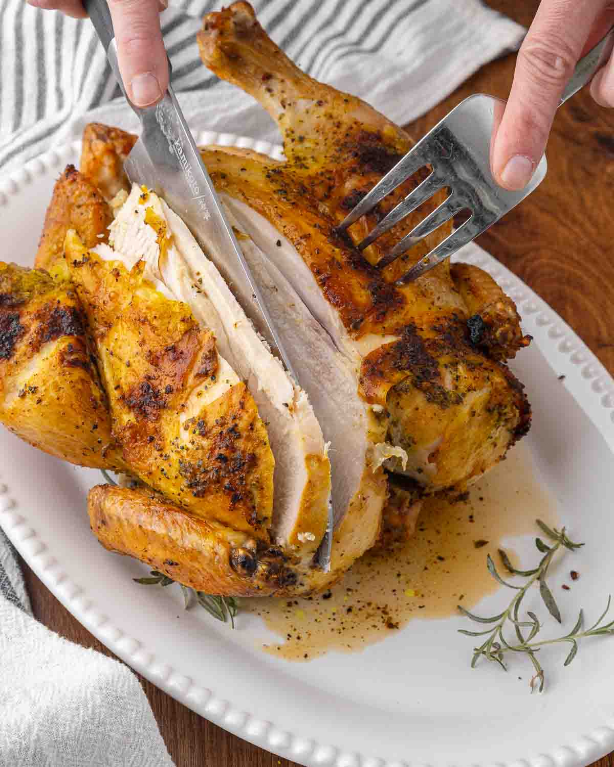 Hands holding a fork and knife, carving a whole roasted chicken.