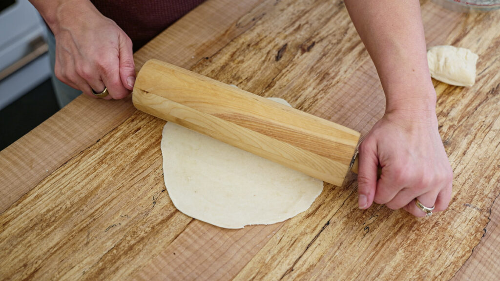 Sourdough tortilla dough being rolled out with a wooden rolling pin.