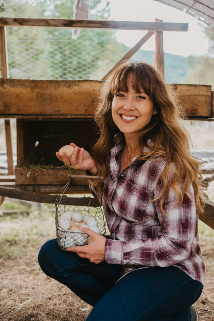 Woman in a mobile chicken tractor holding a wire basket of freshly laid chicken eggs