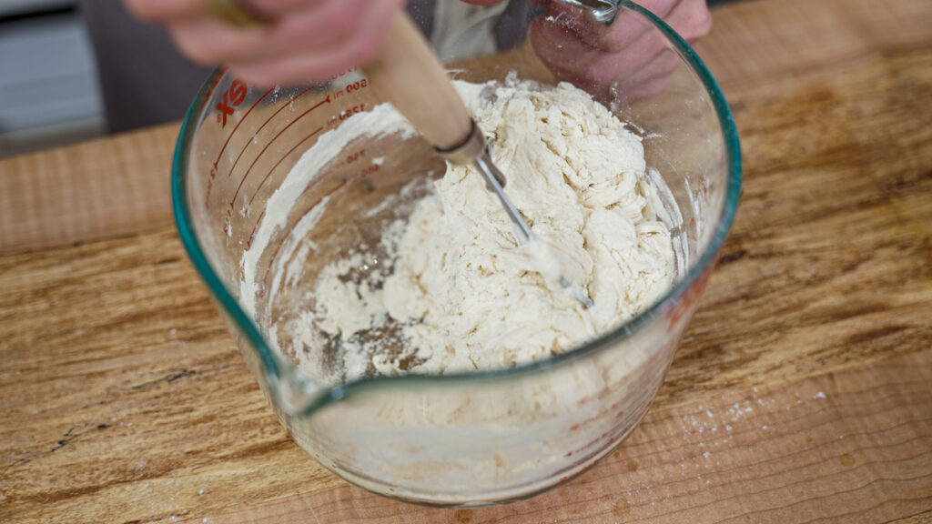 Sourdough tortilla dough being mixed with a Danish dough whisk in a large glass bowl.