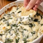 Spinach artichoke dip in a baking dish and a hand with a chip taking a scoop.