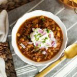 Homemade chili in a white bowl topped with sour cream, onions and cilantro.