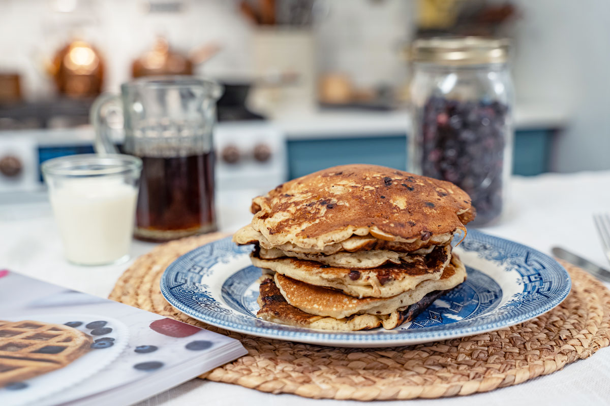 A stack of sourdough pancakes on a blue plate with maple syrup and a jar of freeze dried blueberries in the background.
