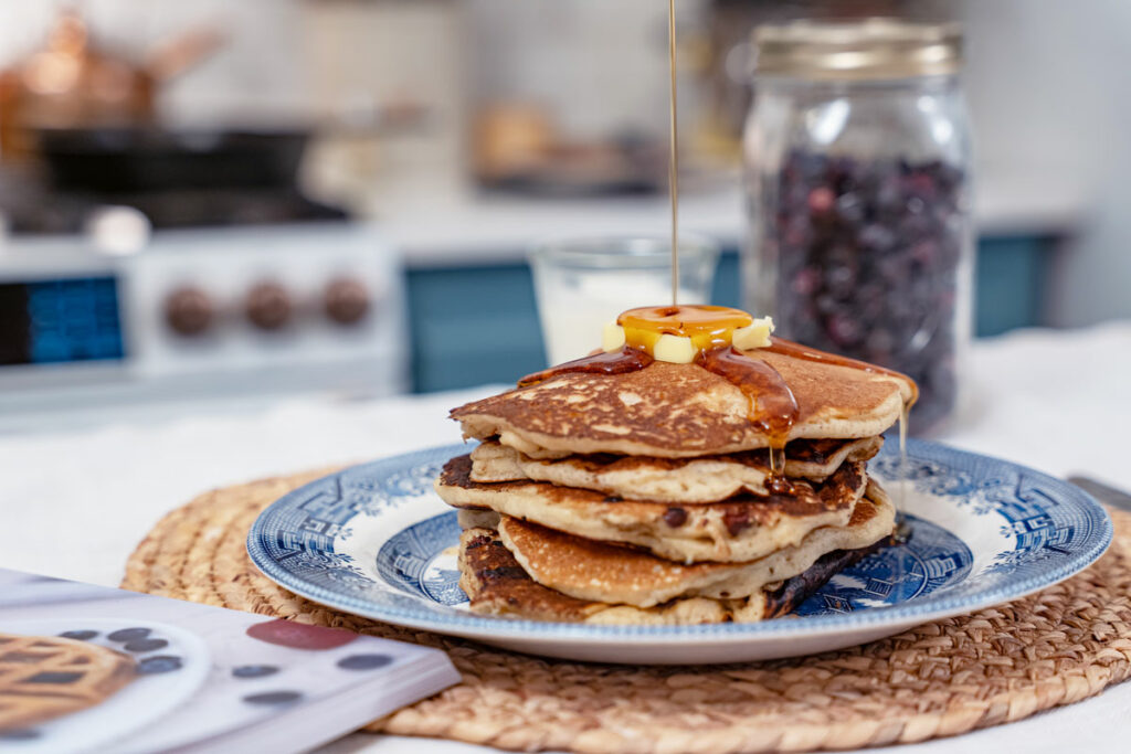 A stack of sourdough pancakes on a blue plate with maple syrup being poured over the top and a jar of freeze dried blueberries in the background.