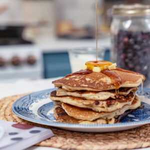 A stack of sourdough pancakes on a blue plate with maple syrup being poured over the top and a jar of freeze dried blueberries in the background.