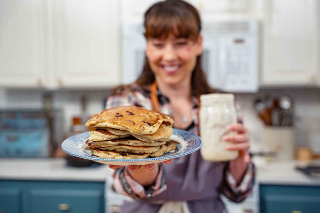 A woman holding up a plate with a stack of pancakes and a jar of sourdough starter in the other hand.