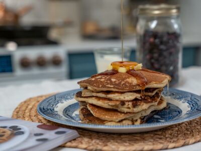 A stack of sourdough pancakes on a blue plate with maple syrup being poured over the top and a jar of freeze dried blueberries in the background.