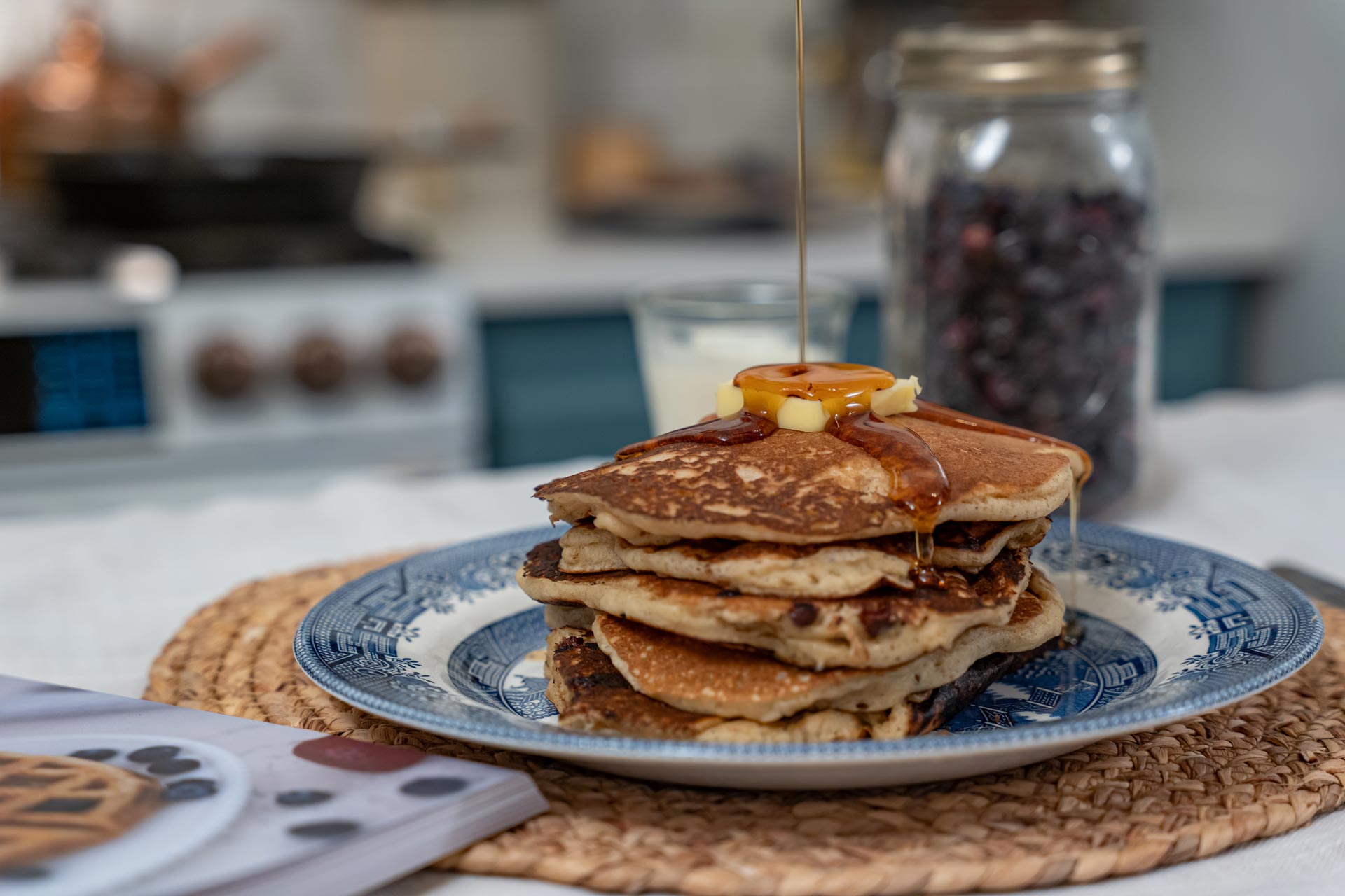 A stack of sourdough pancakes on a blue plate with maple syrup being poured over the top and a jar of freeze dried blueberries in the background.