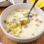 Cream of broccoli cheddar soup in a bowl with a spoon and rolls in the background.