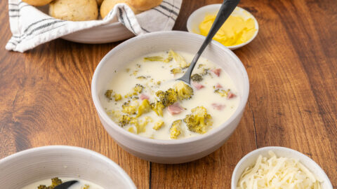 Cream of broccoli cheddar soup in a bowl with a spoon and rolls in the background.