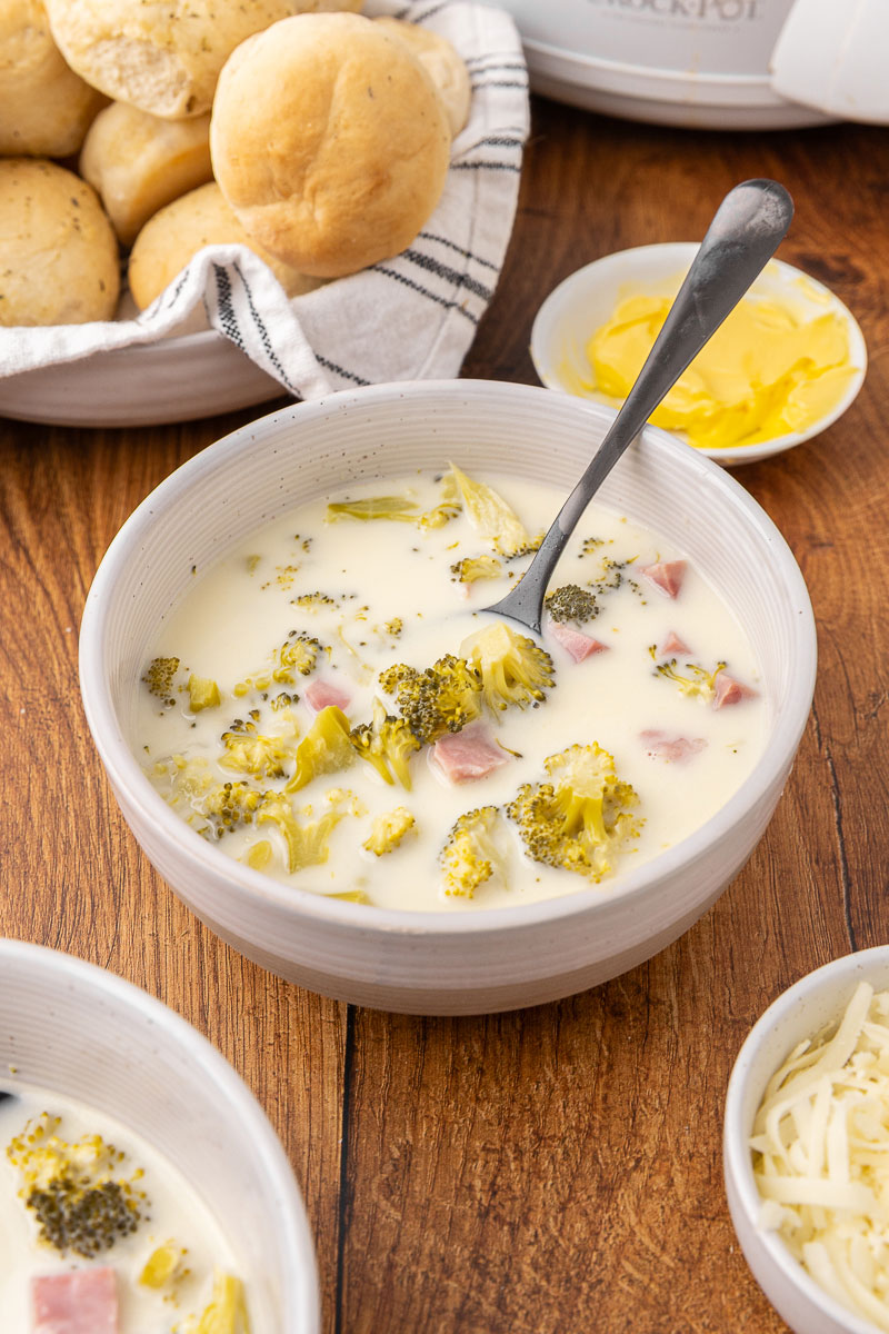 Cream of broccoli cheddar soup in a bowl with a spoon and rolls in the background.