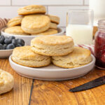 A stack of baked biscuits on a plate with fruit and jam in the background.