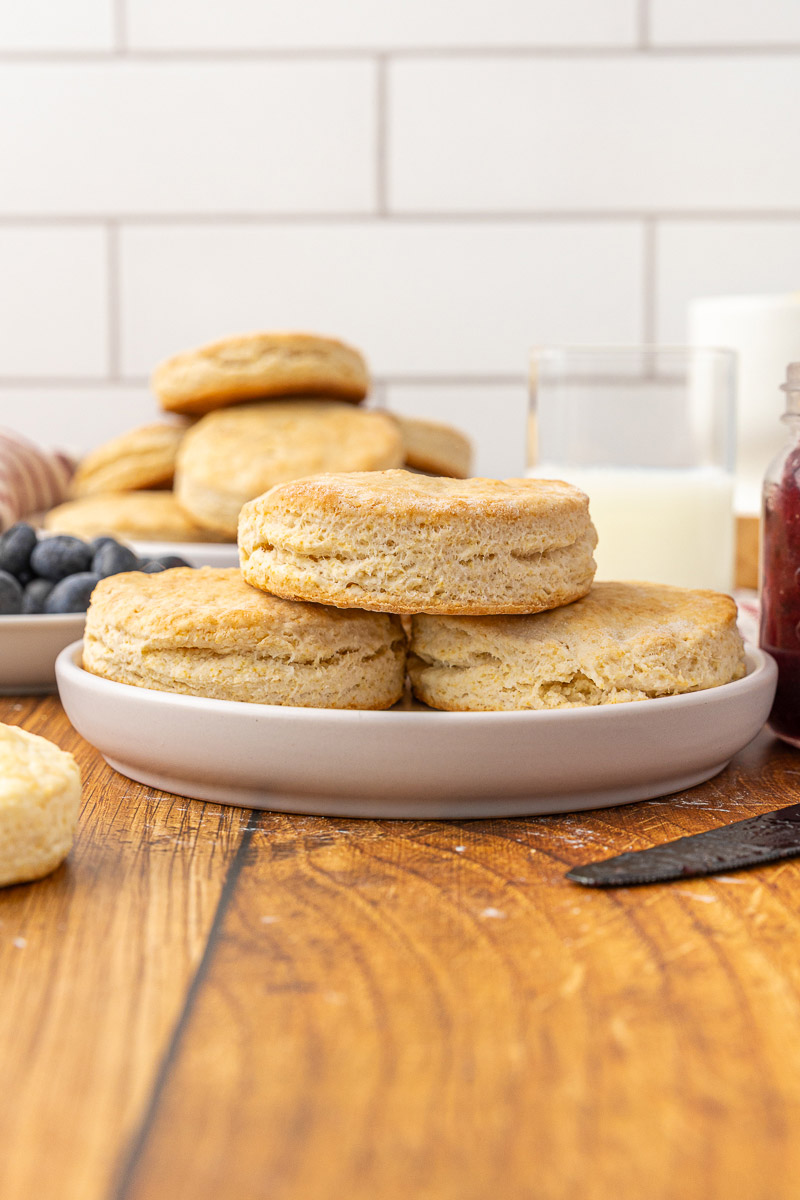 A stack of baked biscuits on a plate with fruit and jam in the background.