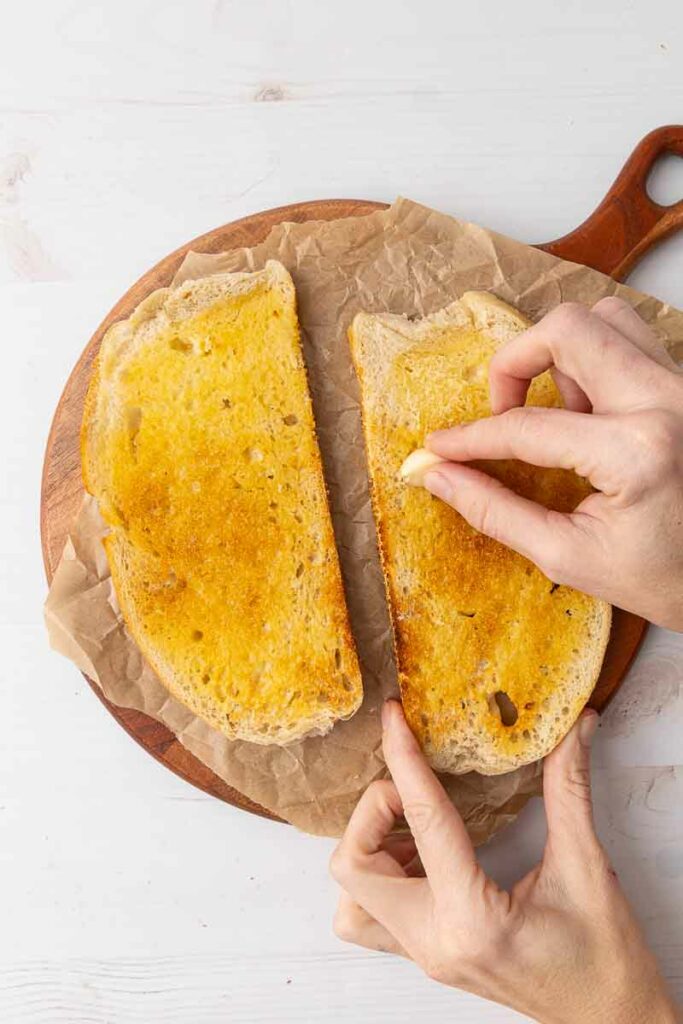 Two pieces of toasted and buttered bread being rubbed with a garlic clove.