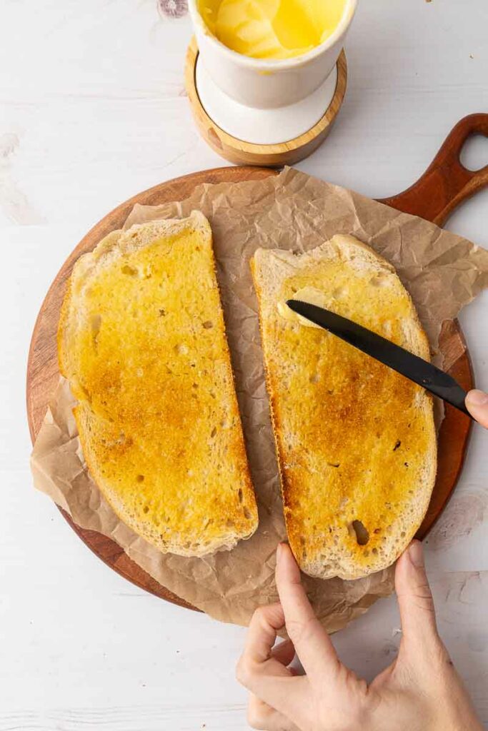 Two pieces of toasted bread being buttered.
