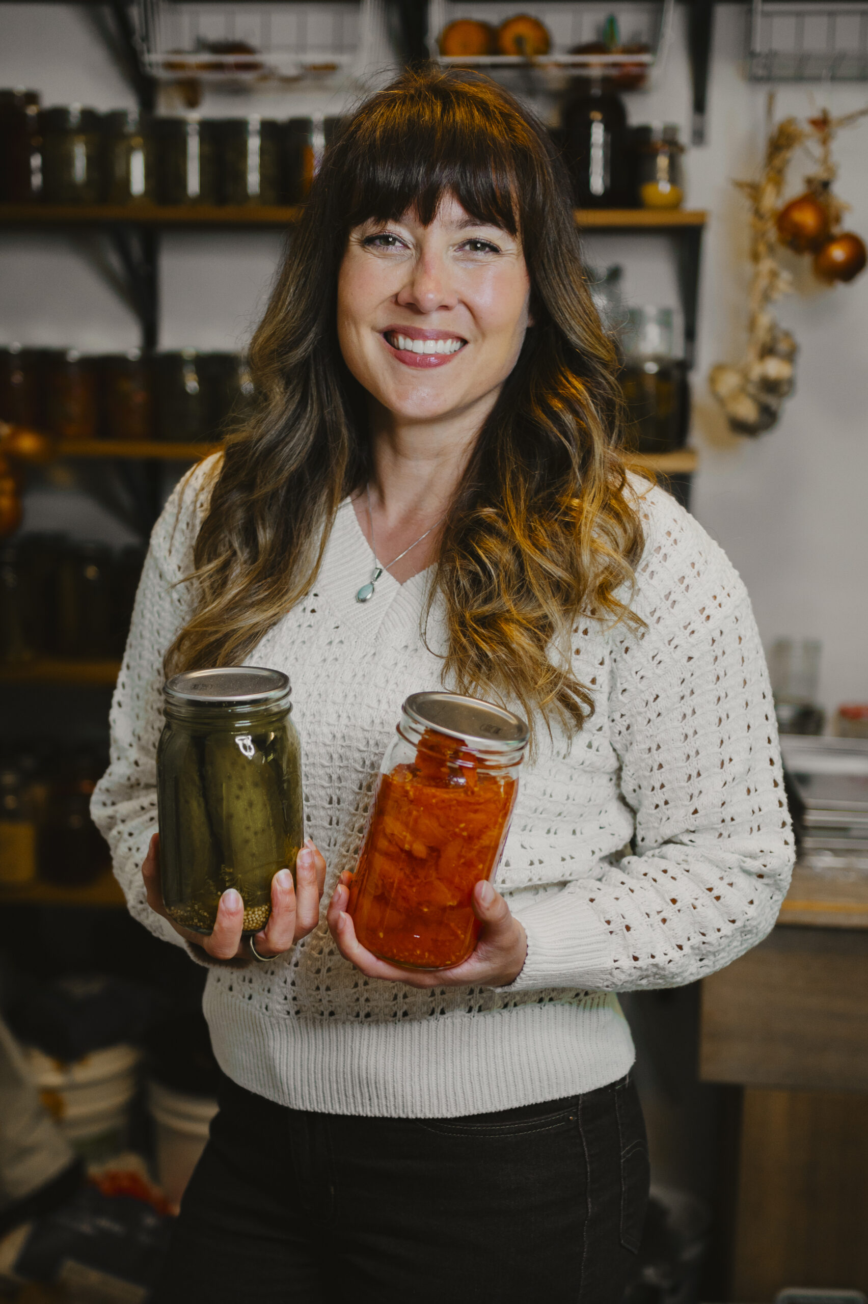 Woman standing infront of rows of home canned food holding a jar of home canned pickles and tomatoes