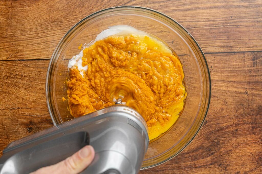 An electric hand mixer whisking together pumpkin, eggs, coconut oil, and sugar.