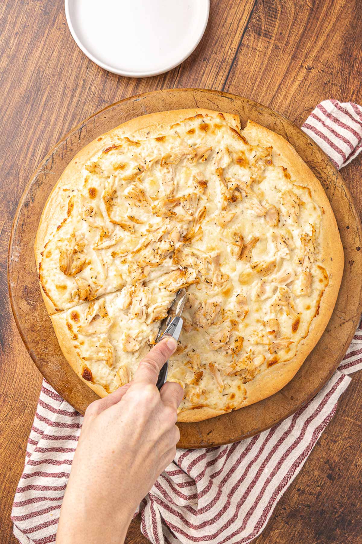 A hand using a pizza cutter to slice chicken Alfredo pizza.