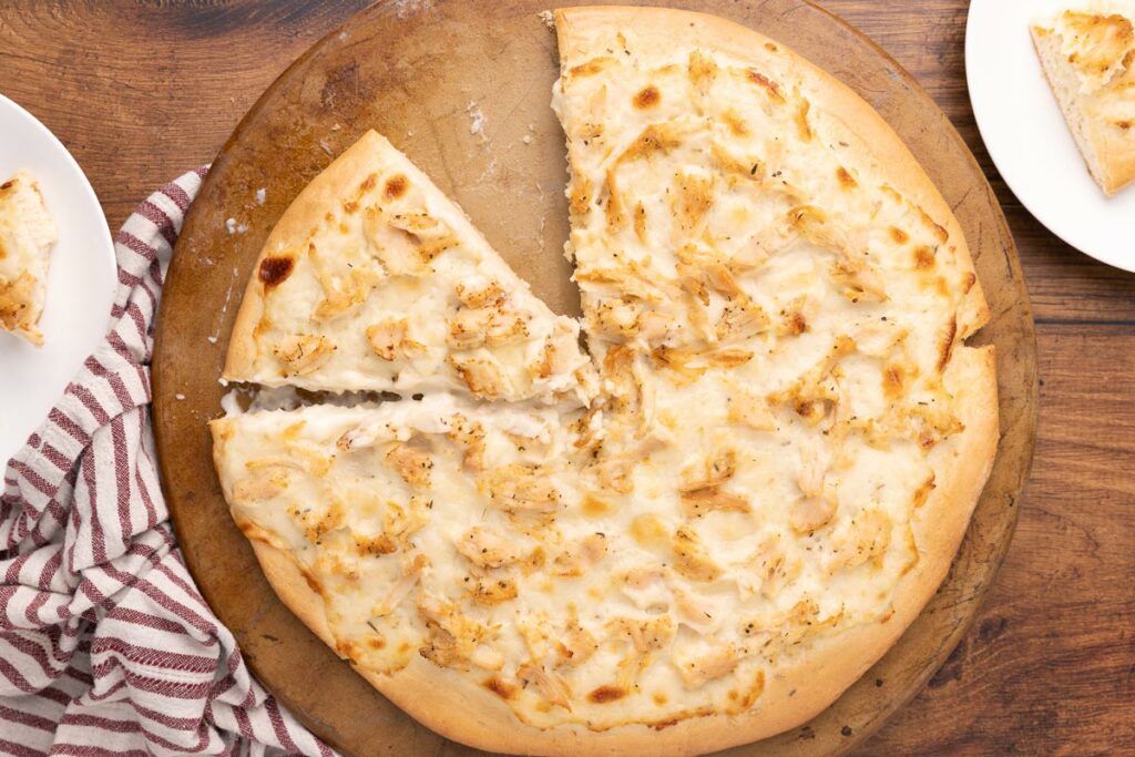 Baked chicken Alfredo pizza on a pizza stone with a slice missing.