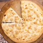Baked chicken Alfredo pizza on a pizza stone with a slice missing.