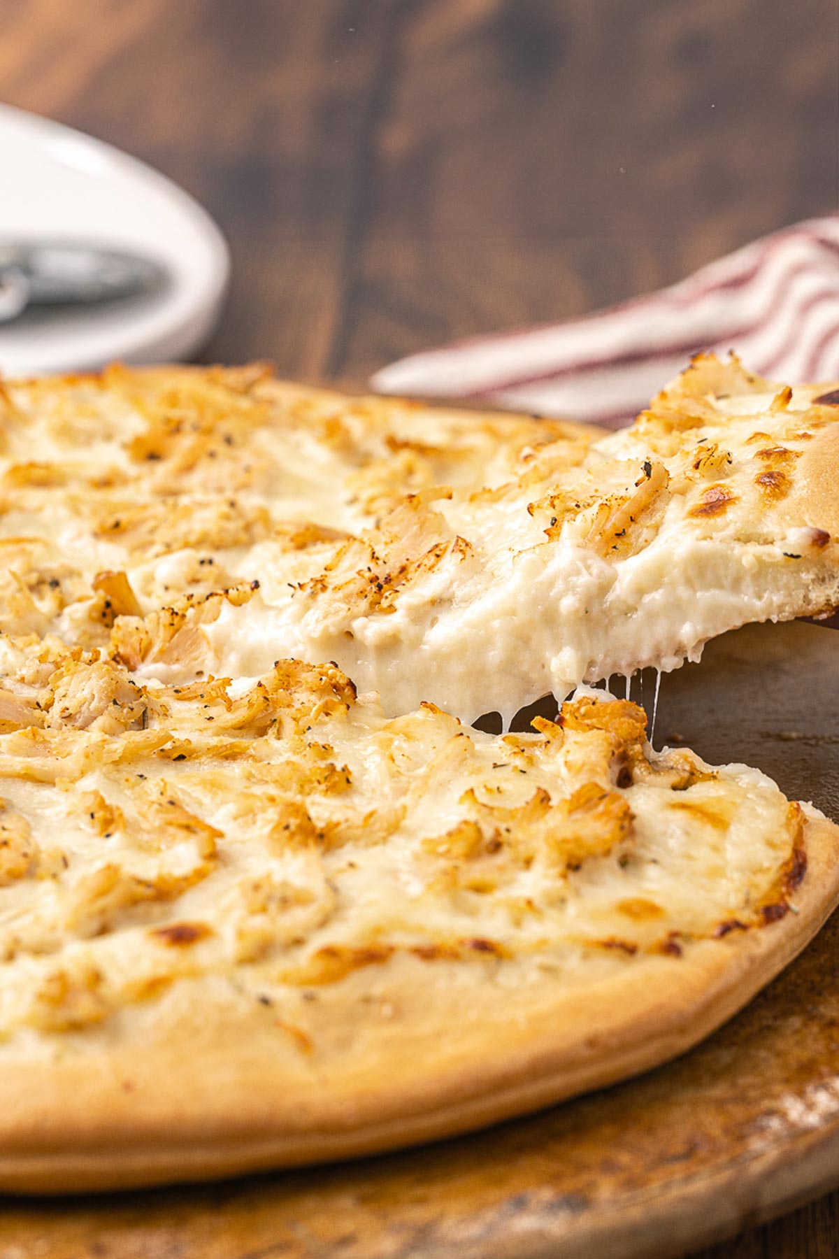 A baked chicken Alfredo pizza on a pizza stone with a hand pulling a piece away.