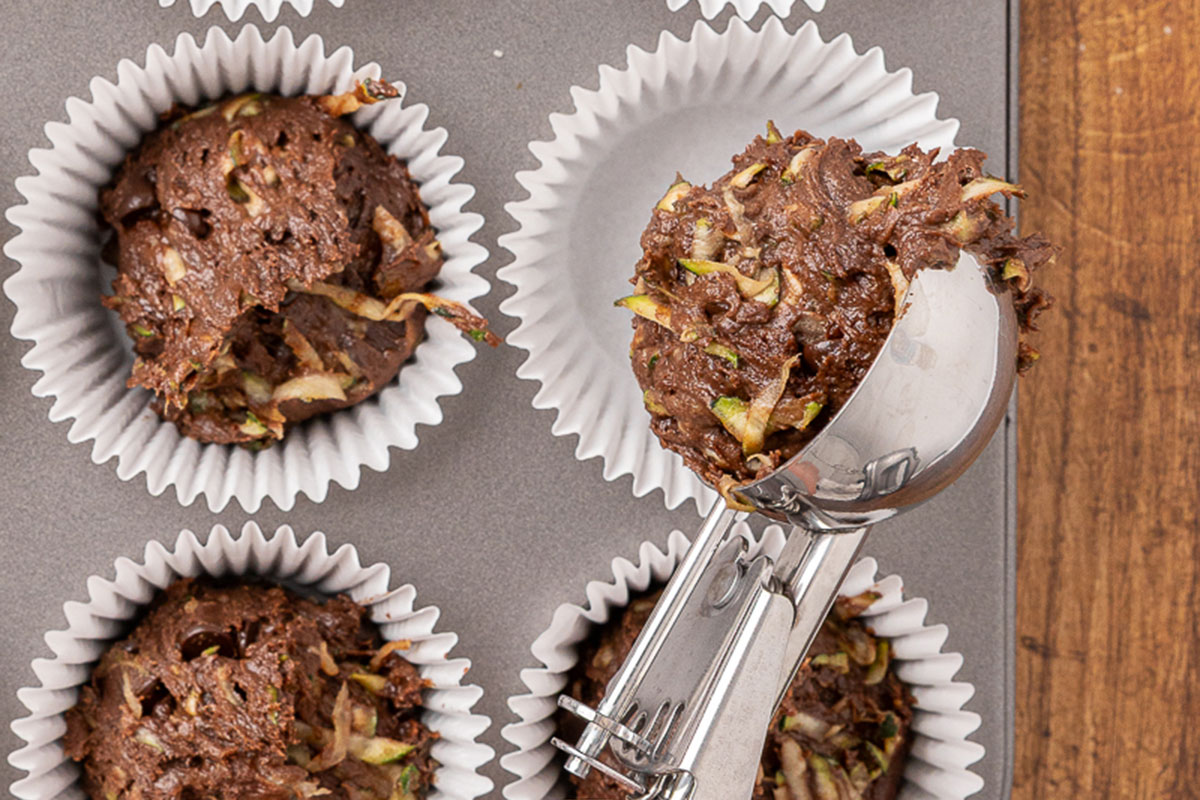 A scoop filling the wells of a muffin tin with muffin batter.