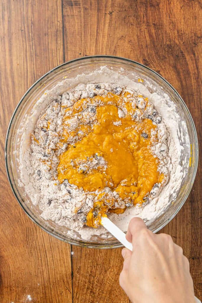 A hand holding a spatula that is folding together the wet and dry ingredients for pumpkin chocolate chip muffins.