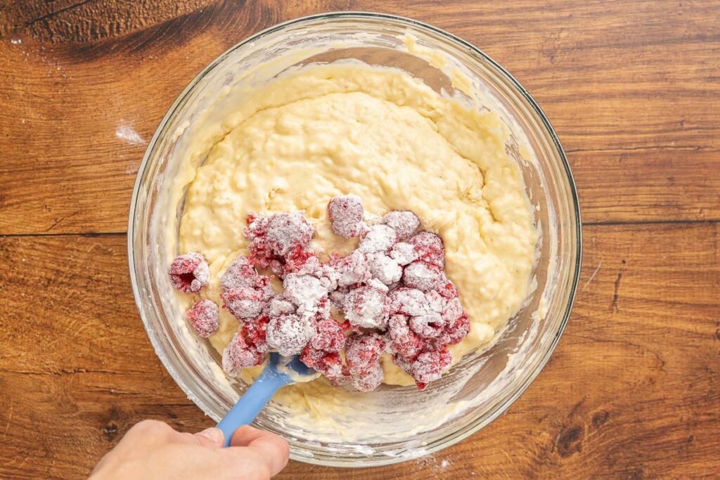 A hand holding a spatula folding raspberries into muffin batter.