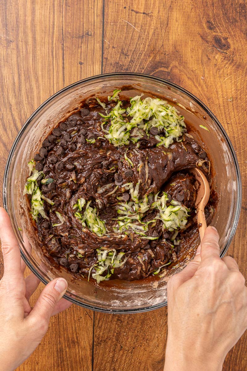 Hands folding shredded zucchini into a bowl of chocolate muffin batter.