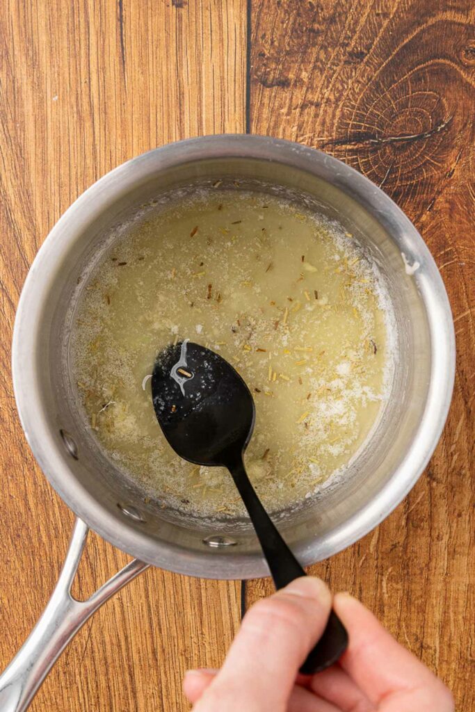 A hand holding a spoon stirring garlic butter melting in a saucepan.