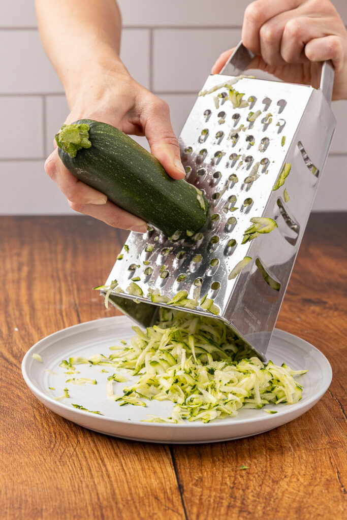 Hands grating a fresh zucchini with a box grater.