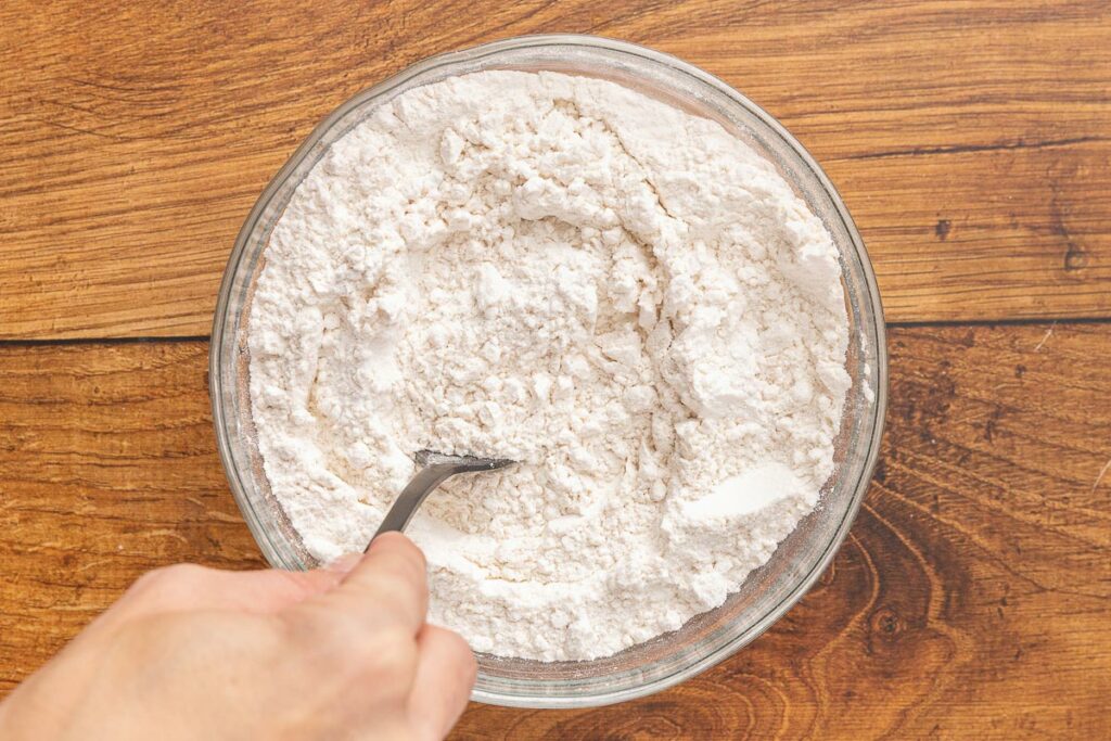 A hand holding a spoon mixing together flour, salt, baking soda, and baking powder.