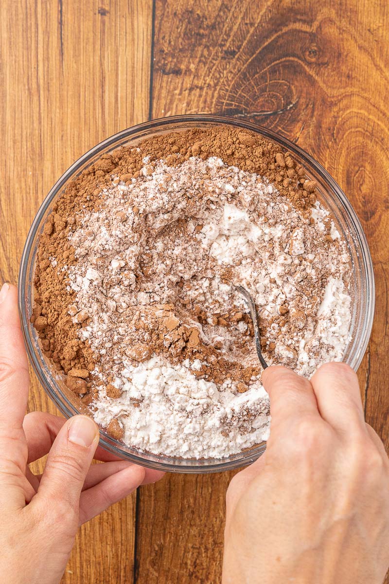 Hands stirring together flour, baking soda, baking powder, salt, and cocoa powder in a bowl.