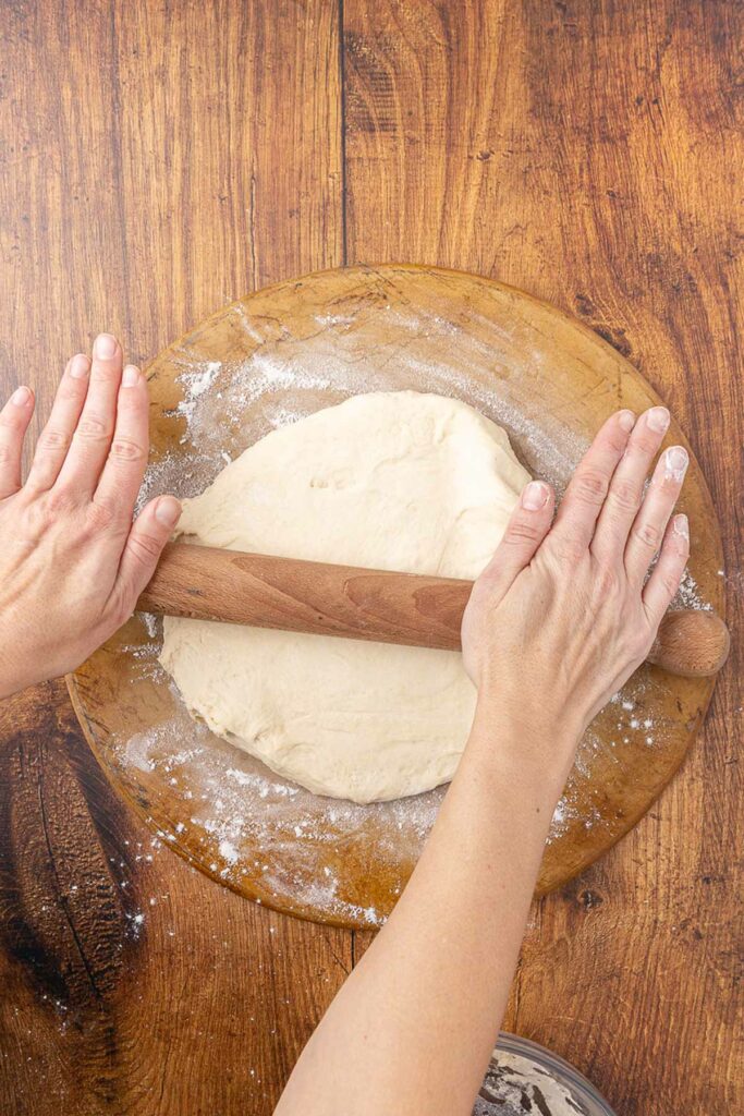 Hands rolling out pizza dough with a wooden rolling pin.