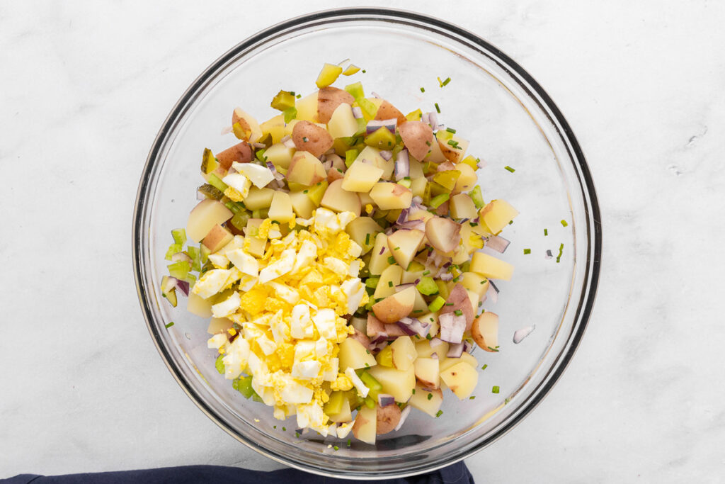 A bowl with chopped pickles, potatoes, hard boiled eggs, and onion ready to mix for potato salad.