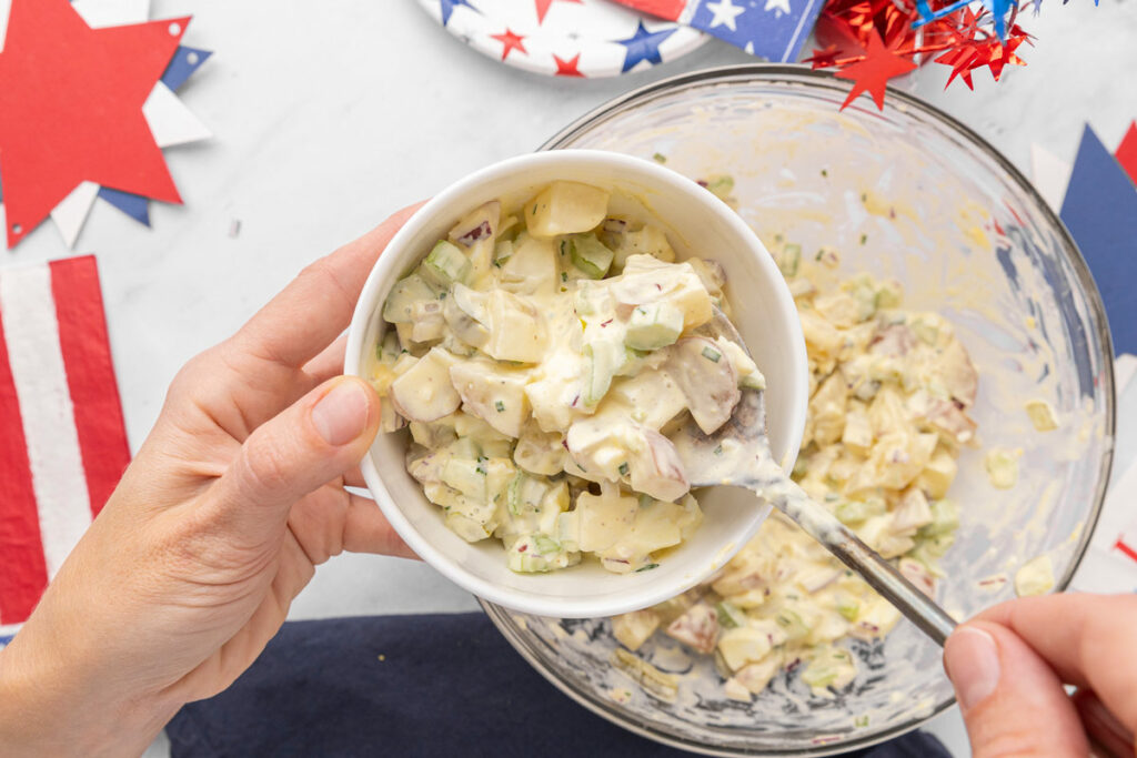 Hands holding a spoon and transferring creamy potato salad from a serving bowl into a storage container.