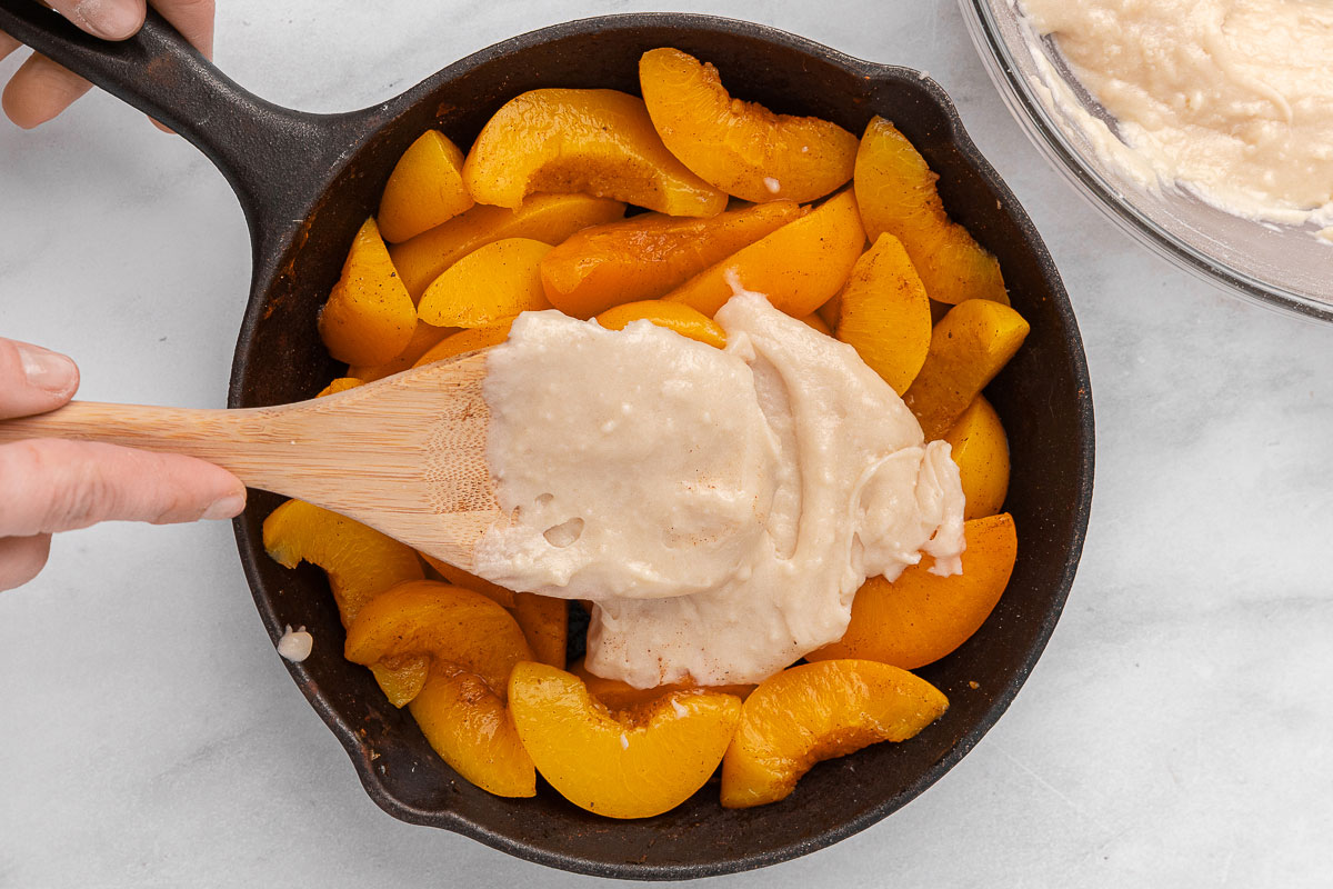 Cobbler topping getting spread over fresh peaches in a cast iron skillet.